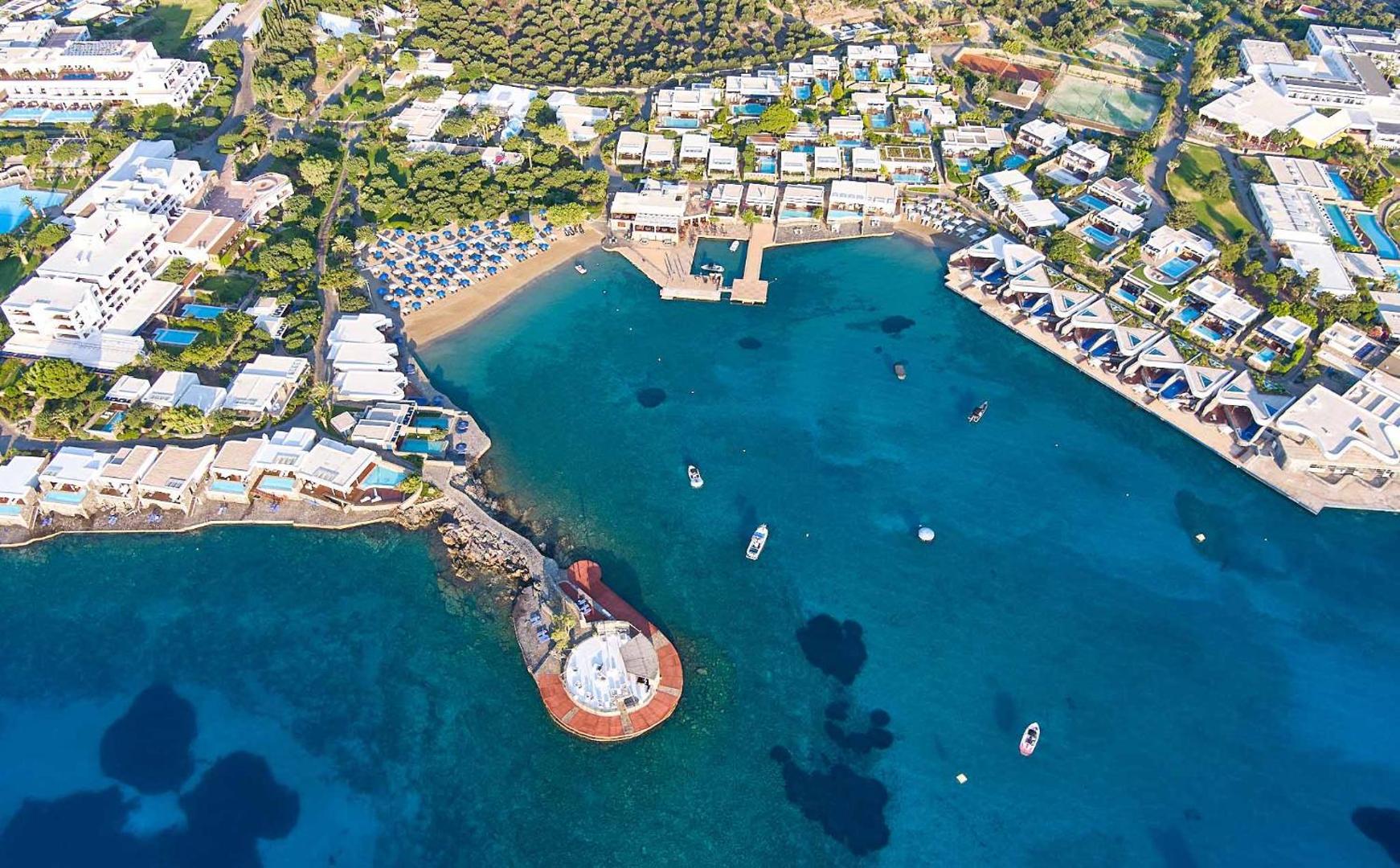 Plašs aerofoto skats uz Elounda Beach Hotel And Villas teritoriju, līci, pludmali un apkārtējiem Krētas kalniem saulainā dienā.
