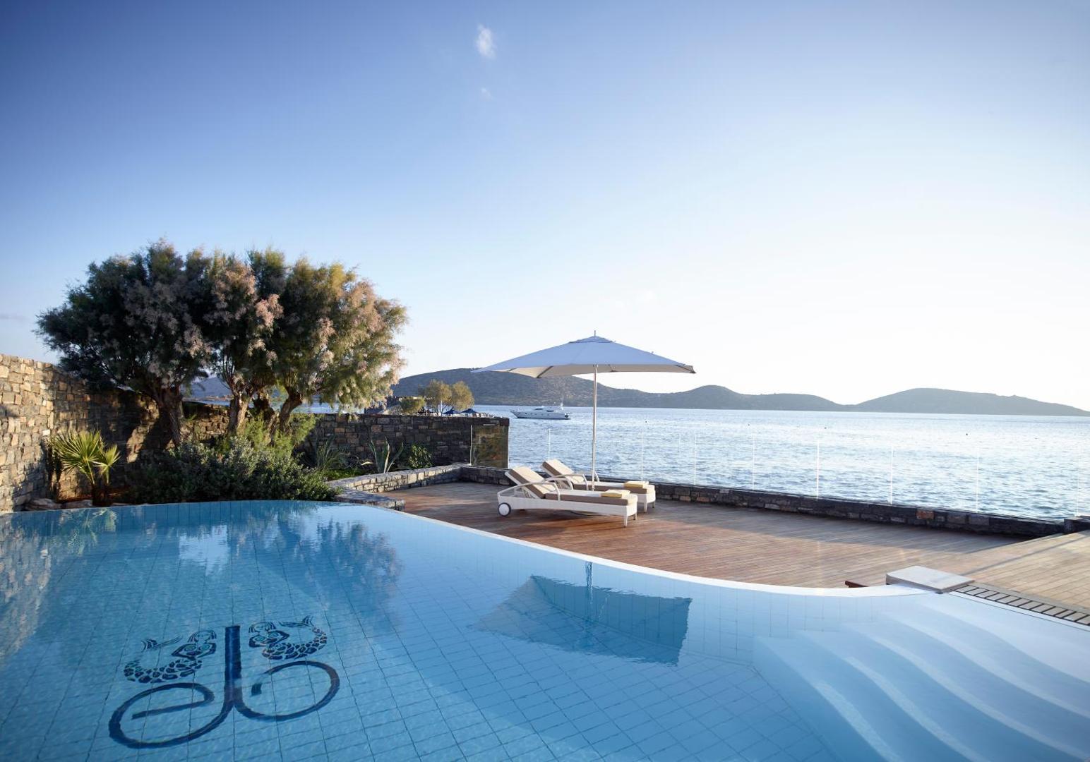Elounda Beach Hotel And Villas baseins ar pārplūdes malu, koka klājs un skats uz mierīgo jūras līci