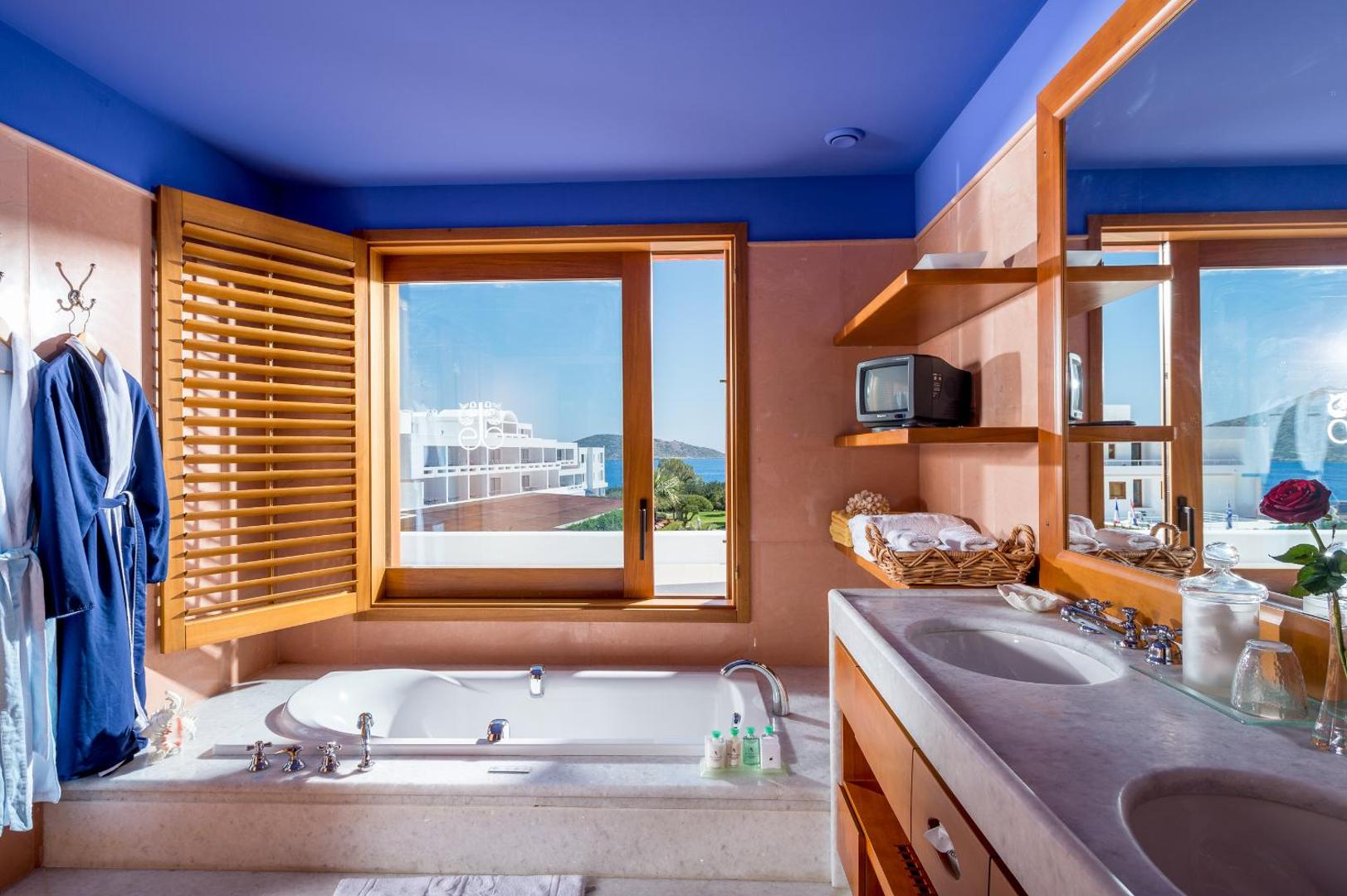 Elounda Beach Hotel luksusa numura vannas istaba ar marmora apdari, iebūvētu vannu un skatu pa logu uz kūrorta teritoriju