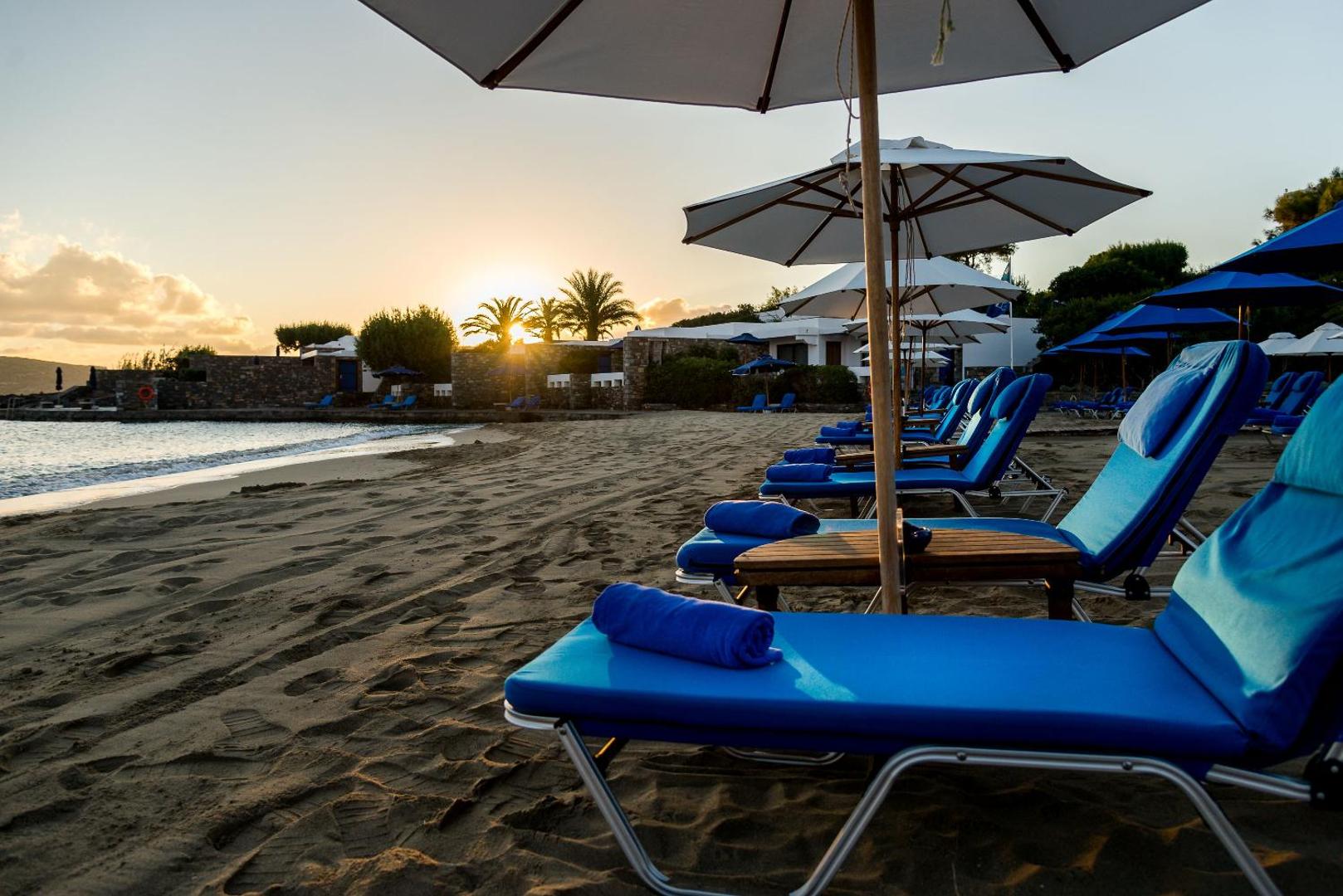 Elounda Beach Hotel And Villas privātā pludmale saulrietā Krētā — zili sauļošanās krēsli un balti saulessargi smilšainajā krastā