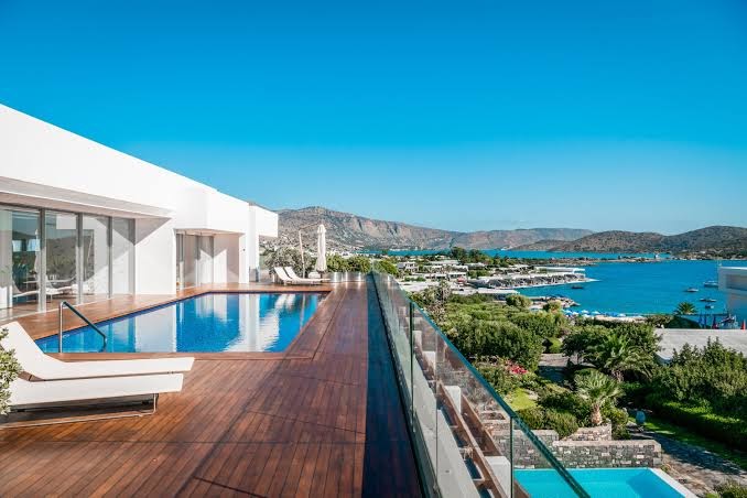 Elounda Beach Hotel And Villas terase ar koka klāju, sauļošanās krēslu un baseinu ar skatu uz līci