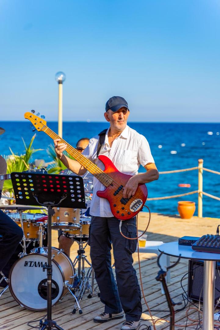 Dzīvās mūzikas priekšnesums Ulusoy Kemer Holiday Club pludmales piestātnē — basģitārists uzstājas uz koka klāja pie Vidusjūras