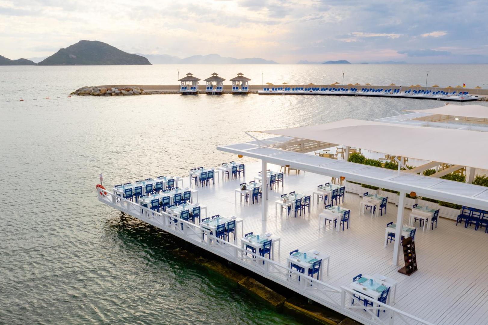 La Blanche Resort & Spa restorāna terase ar skatu uz Egejas jūru un kalniem — balti klāti galdi, saulains laiks un mols fonā