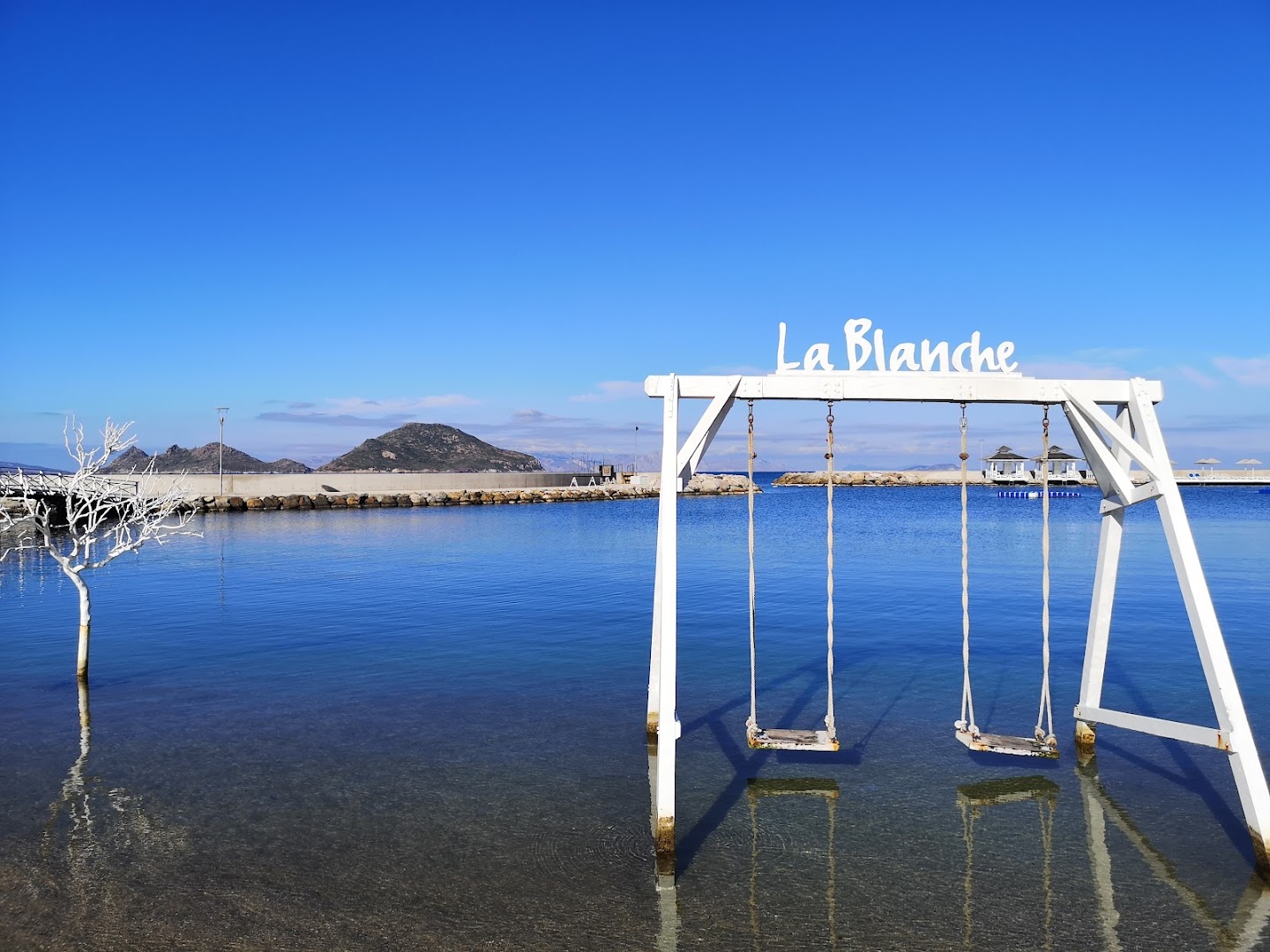 La Blanche Resort & Spa dekoratīvās šūpoles jūrā ar viesnīcas nosaukumu — dzidrs ūdens, zilas debesis un kalni tālumā