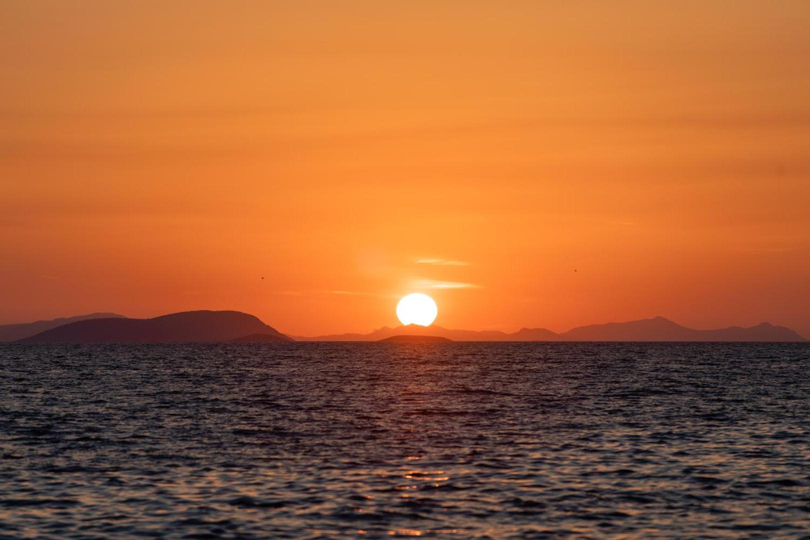 Tuvplāns ar saulrietu virs Egejas jūras horizonta un kalnu siluetiem netālu no La Blanche Resort & Spa kūrorta