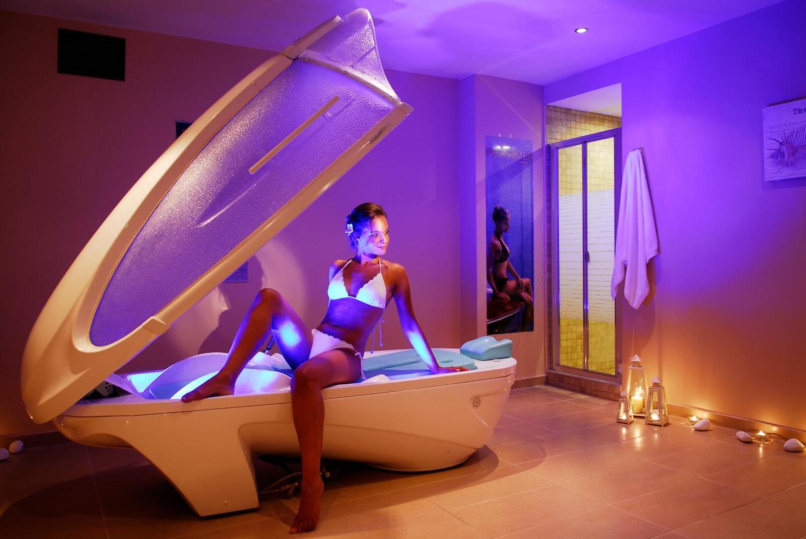 Sieviete peldkostīmā sēž modernā spa kapsulā ar violetu apgaismojumu.