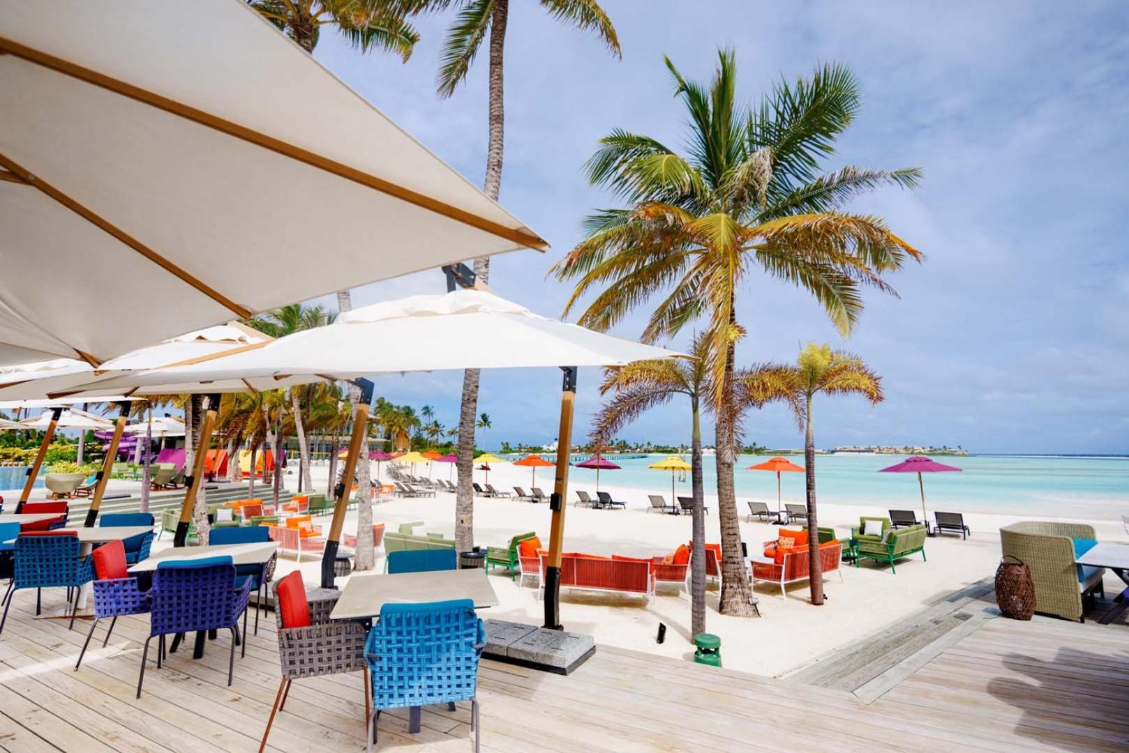 Hard Rock Hotel Maldives pludmales bārs un atpūtas zona ar krāsainiem krēsliem, saulessargiem un skatu uz tirkīza zilo lagūnu