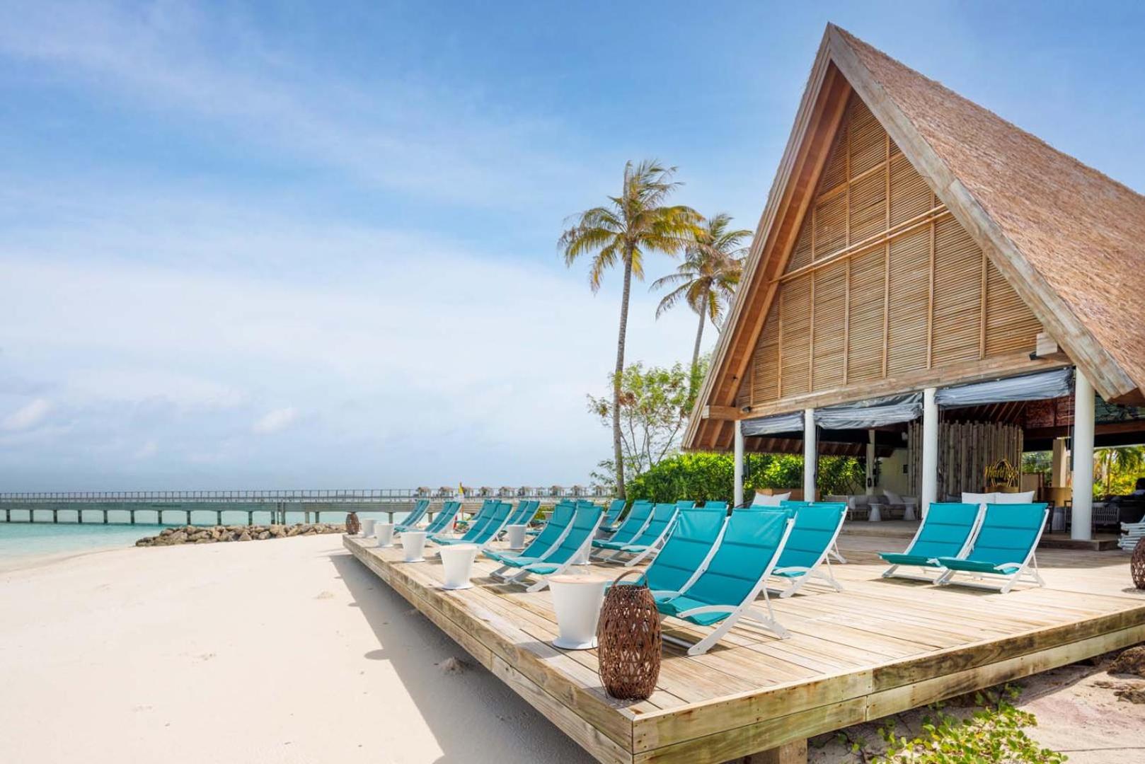 Hard Rock Hotel Maldives pludmales klubs ar tirkīza krāsas zviļņiem uz koka terases pie baltu smilšu pludmales un palmu fona