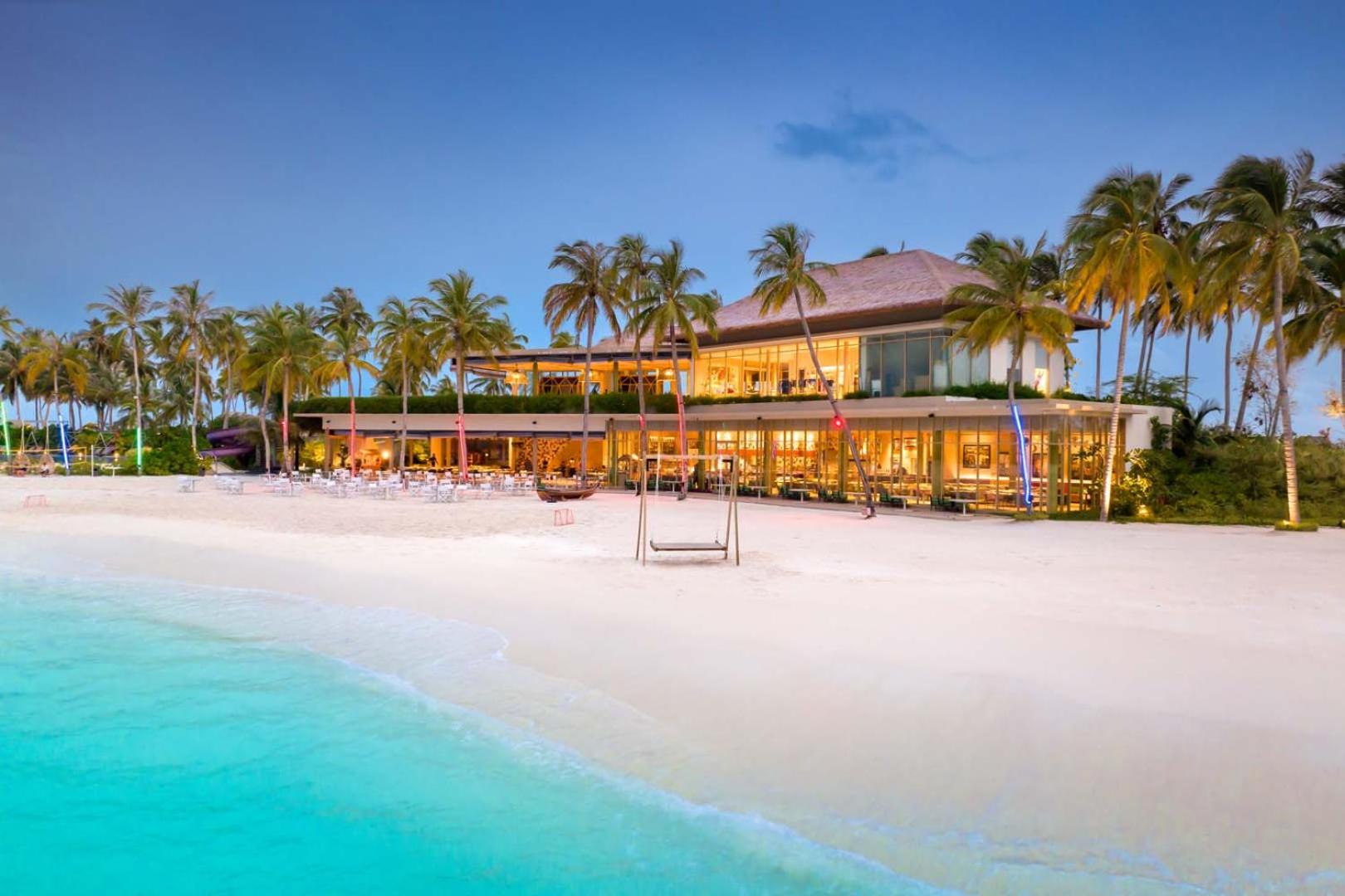 Hard Rock Hotel Maldives pludmales restorāns vakarā — apgaismota ēka pie baltu smilšu pludmales un tirkīza ūdens Maldivu salās
