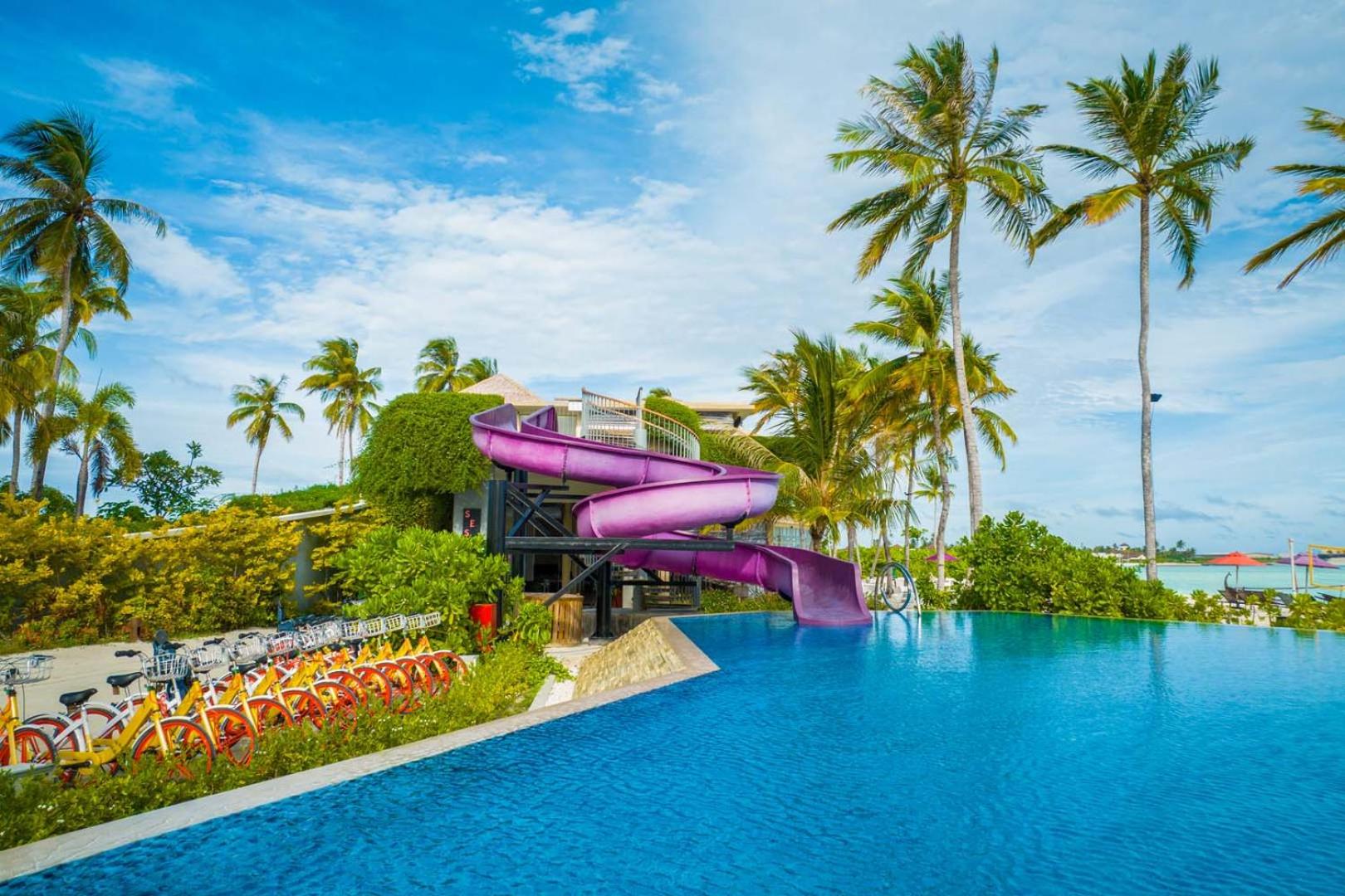 Hard Rock Hotel Maldives āra baseins ar violetu ūdens slidkalniņu, palmām un velosipēdu nomas punktu Maldīvu kūrortā