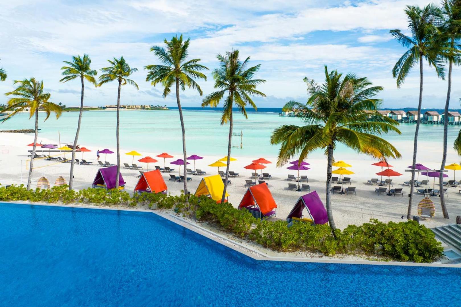 Hard Rock Hotel Maldives skats no augšas uz zilo baseinu, balto smilšu pludmali ar krāsainām teltīm un palmām Maldīvu salās