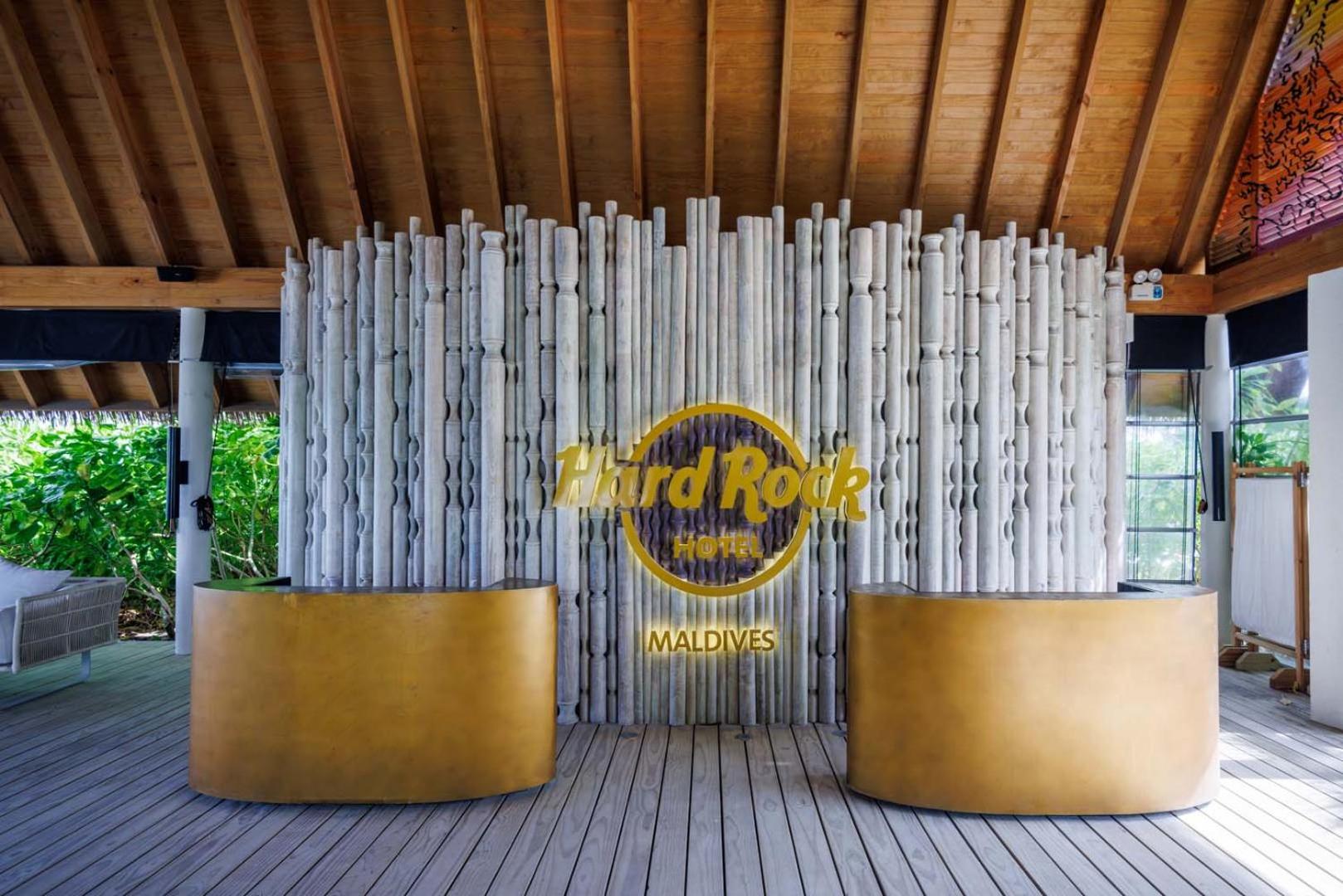 Hard Rock Hotel Maldives reģistratūras zona ar zelta krāsas letēm un dekoratīvu koka elementu sienu tropu stilā