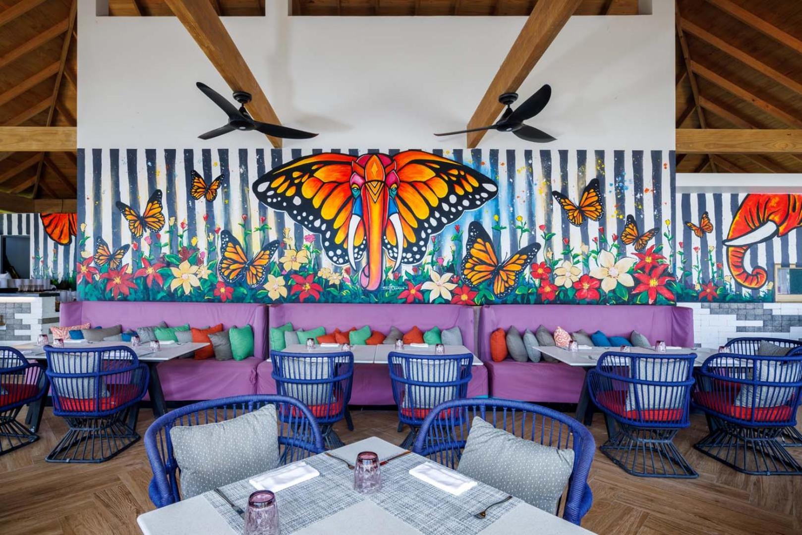 Hard Rock Hotel Maldives restorāna interjers ar košu sienas gleznojumu, kurā attēlots zilonis un tauriņi, ziliem krēsliem un violetiem dīvāniem