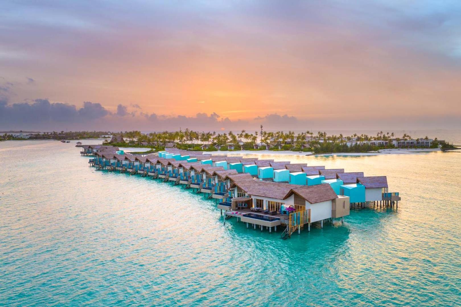 Hard Rock Hotel Maldives ūdens bungalo rinda saulrietā — koka laipas un villas virs okeāna Maldivu salu kūrortā