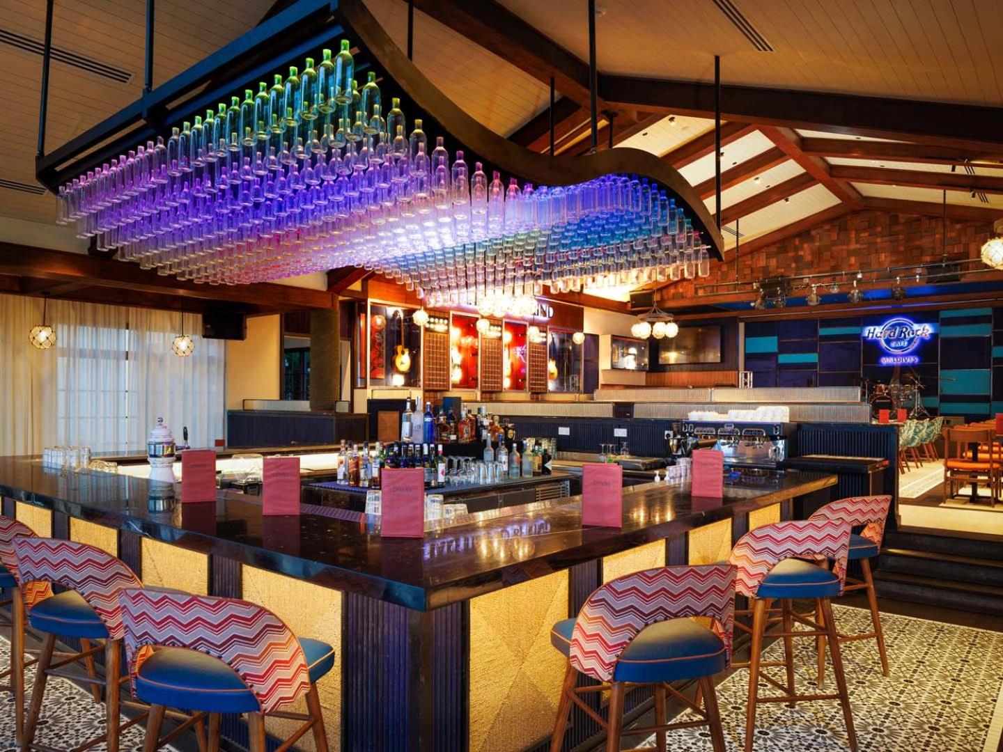 Hard Rock Cafe Maldives bāra lete ar krāsainu pudeļu instalāciju pie griestiem un stilīgiem bāra krēsliem — vakara izklaides vieta