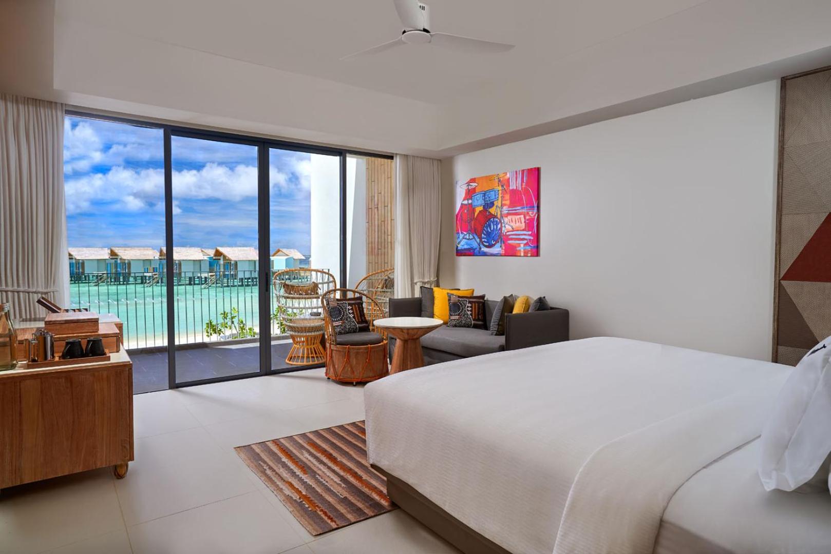 Hard Rock Hotel Maldives numurs ar skatu uz ūdens bungalo ciematu, modernu mēbelējumu un plašu stikloto sienu uz balkonu.