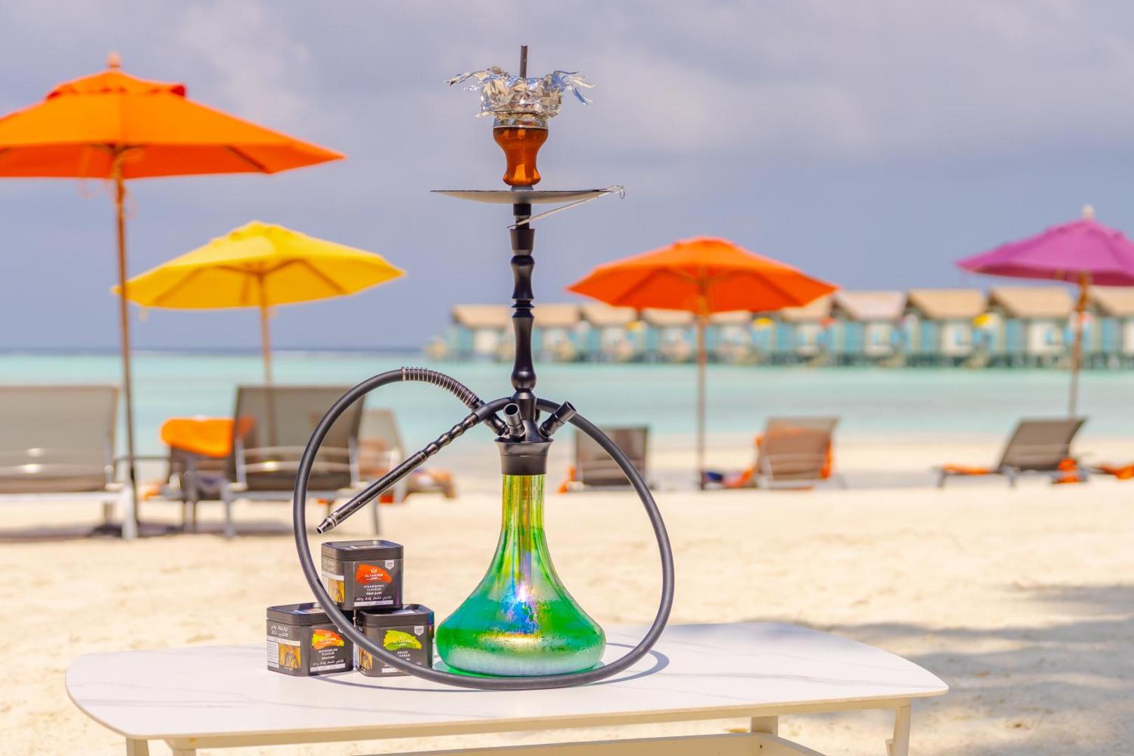 Ūdenspīpe uz galda Hard Rock Hotel Maldives pludmalē, fonā krāsaini saulessargi, okeāns un bungalo uz ūdens