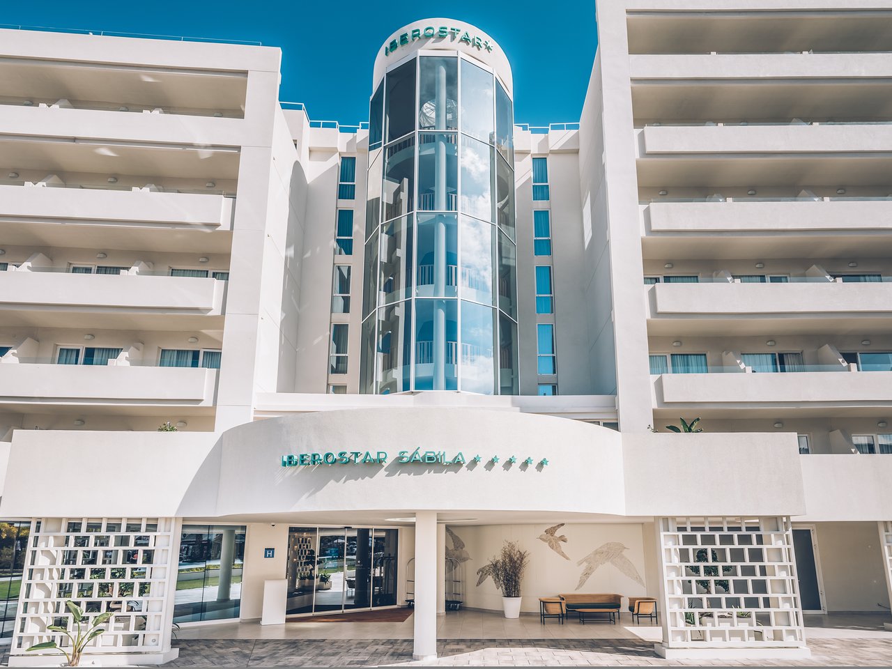 Iberostar Selection Sábila galvenā ieeja Tenerifē — moderna balta arhitektūra ar stikla liftu un viesnīcas logo pie ieejas