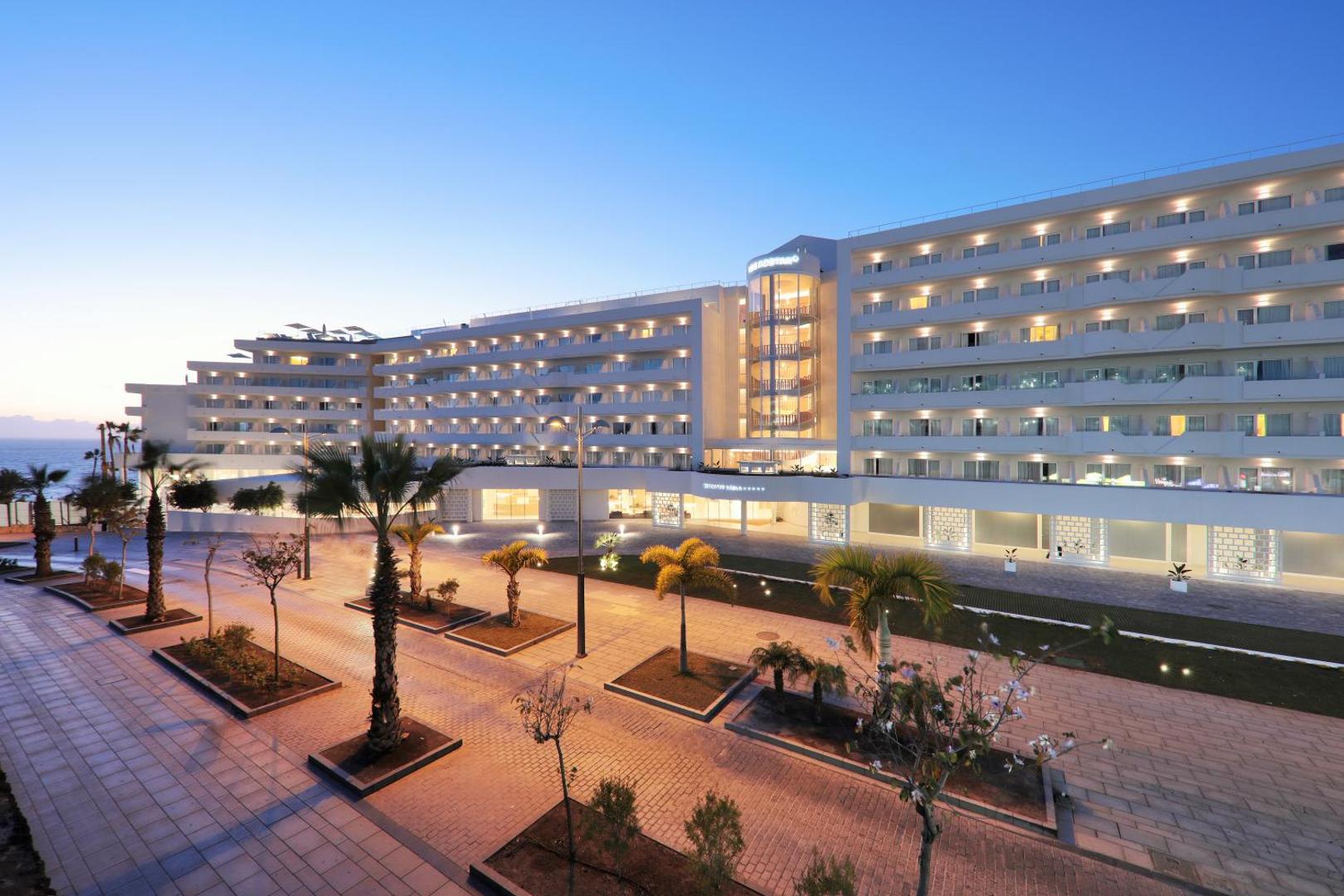 Iberostar Selection Sábila viesnīcas fasāde vakarā Tenerifē — apgaismota ēka ar balkoniem, palmām un bruģētu piebraucamo ceļu saulrieta laikā