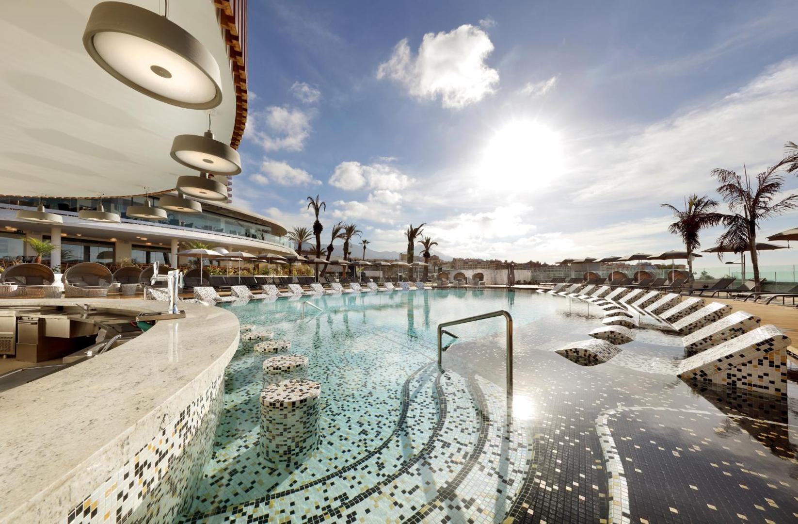 Hard Rock Hotel Tenerife modernais baseina bārs ar mozaīkas flīžu krēsliem ūdenī un saulainu terasi piekrastē