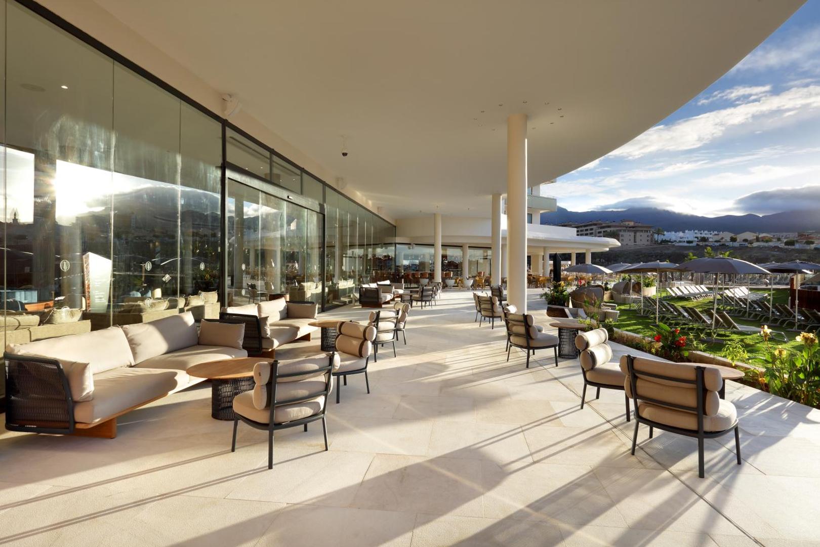 Hard Rock Hotel Tenerife āra terase ar ērtiem dīvāniem un skatu uz kalniem un baseina zonu saulainā dienā