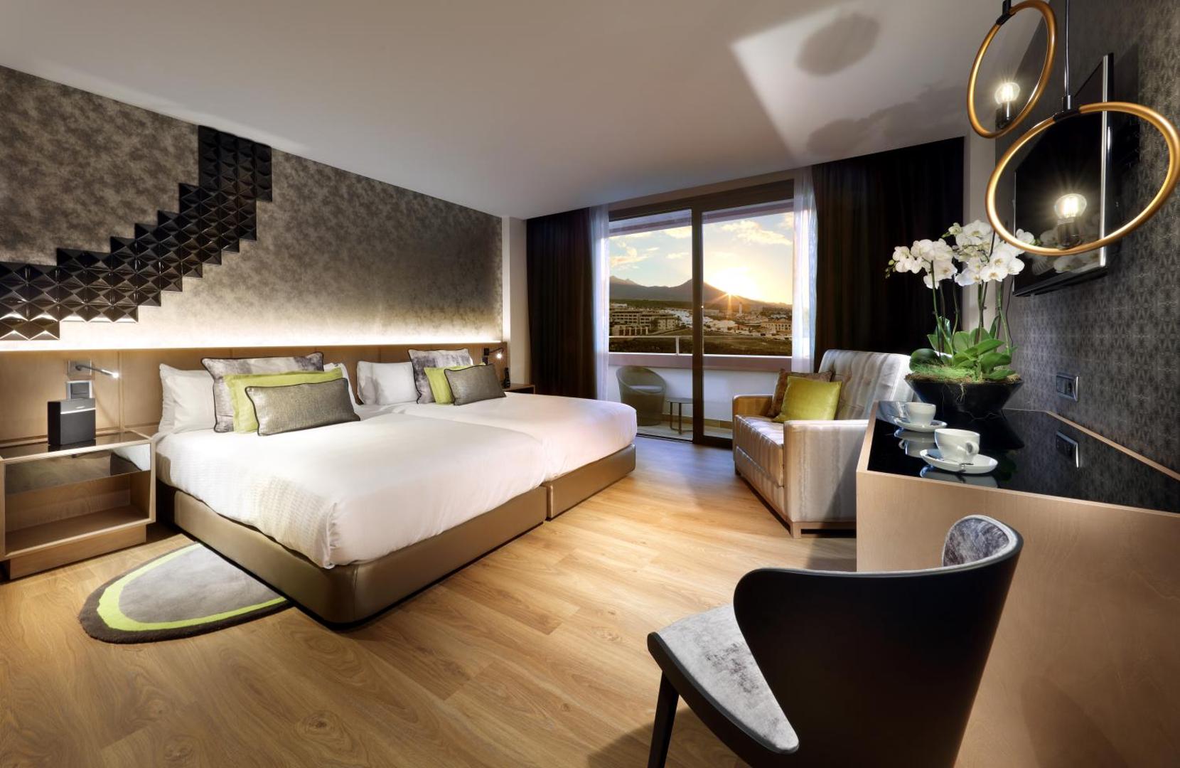 Hard Rock Hotel Tenerife moderns numurs ar divām gultām, atpūtas zonu un balkonu, no kura paveras skats uz kalniem saulrietā.