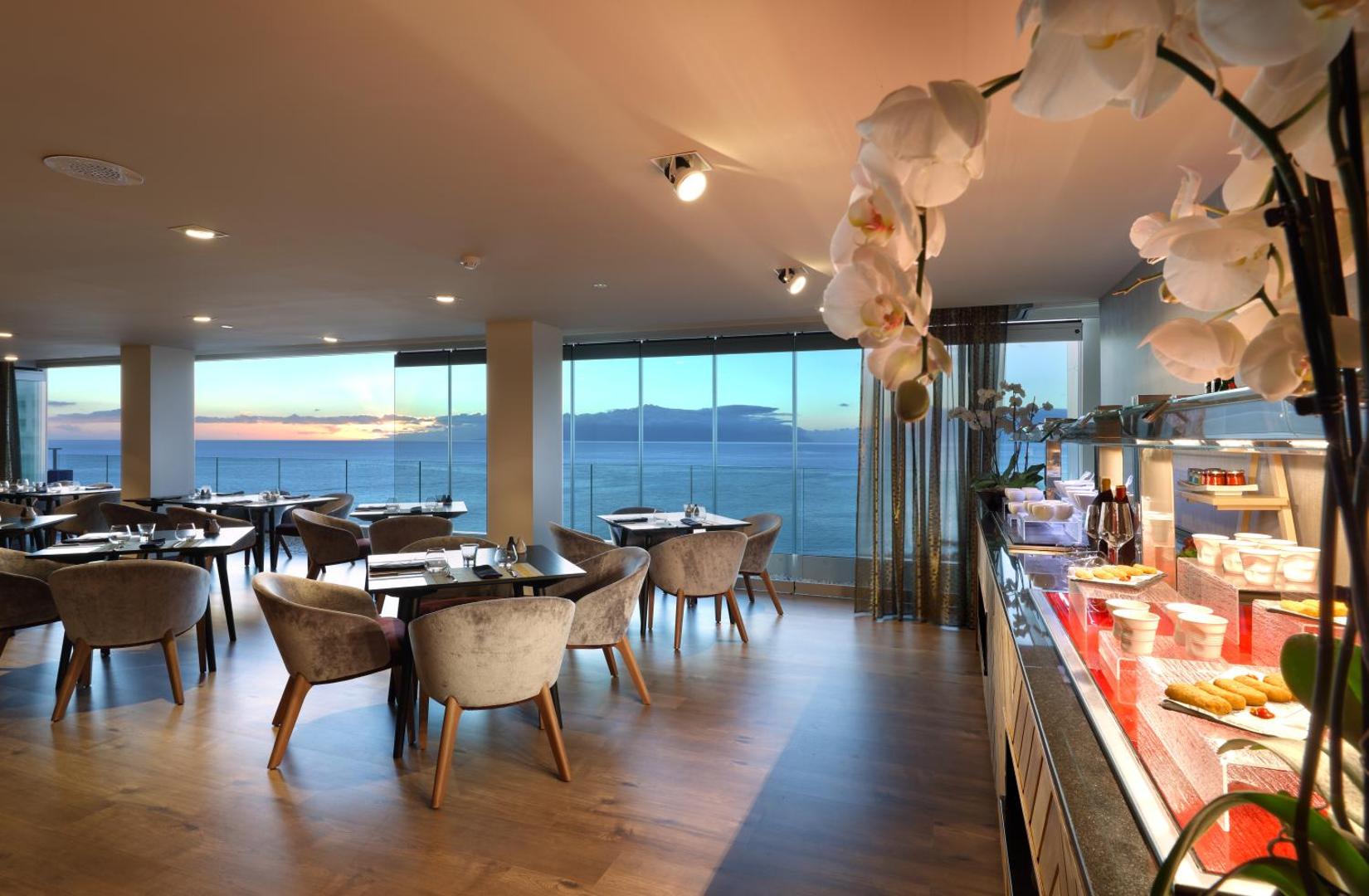 Hard Rock Hotel Tenerife restorāna interjers ar bufetes zonu un panorāmas logiem uz okeānu saulrieta stundā