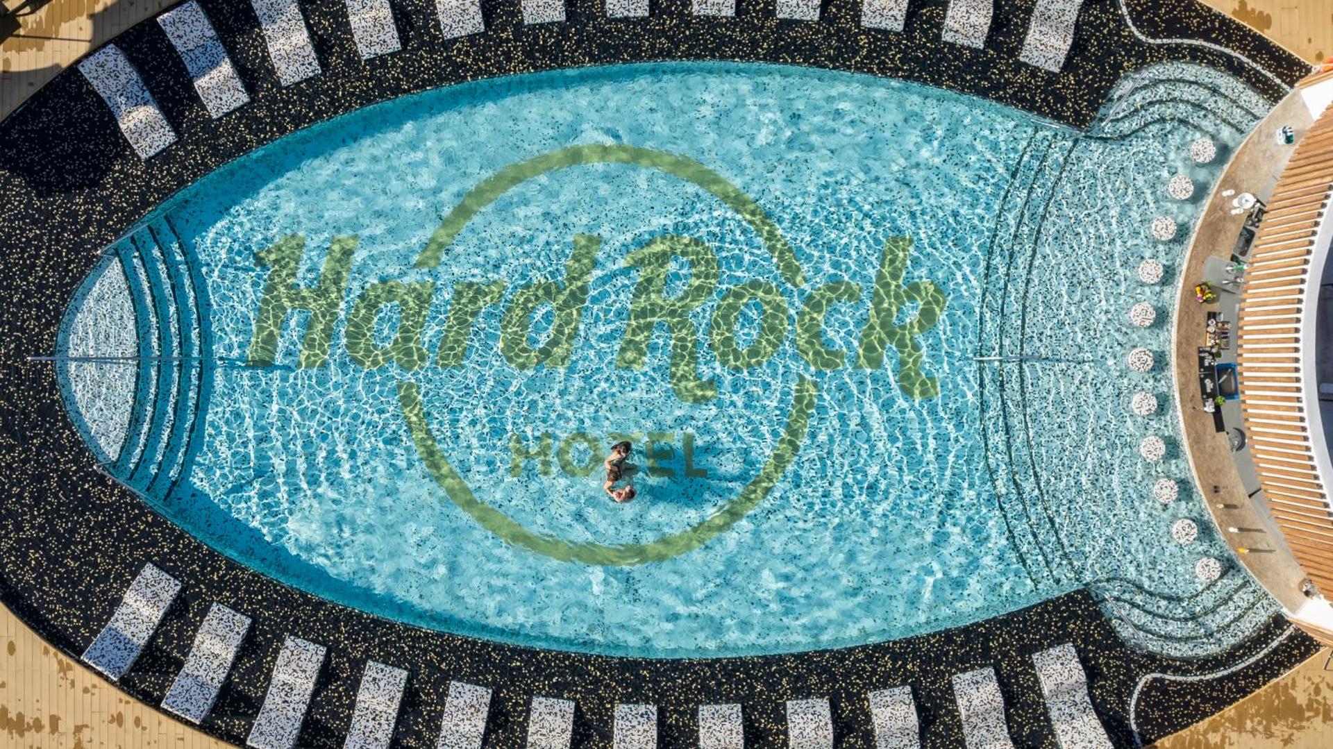 Skats no augšas uz Hard Rock Hotel Tenerife baseinu ar viesnīcas logo apakšā, ko ieskauj stilīga sauļošanās zona.