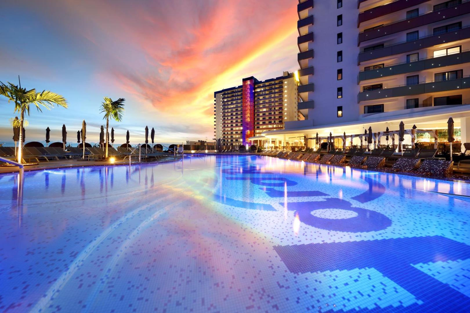 Hard Rock Hotel Tenerife āra baseins un viesnīcas ēkas saulrieta laikā ar krāsainām debesīm un izgaismotām palmām.