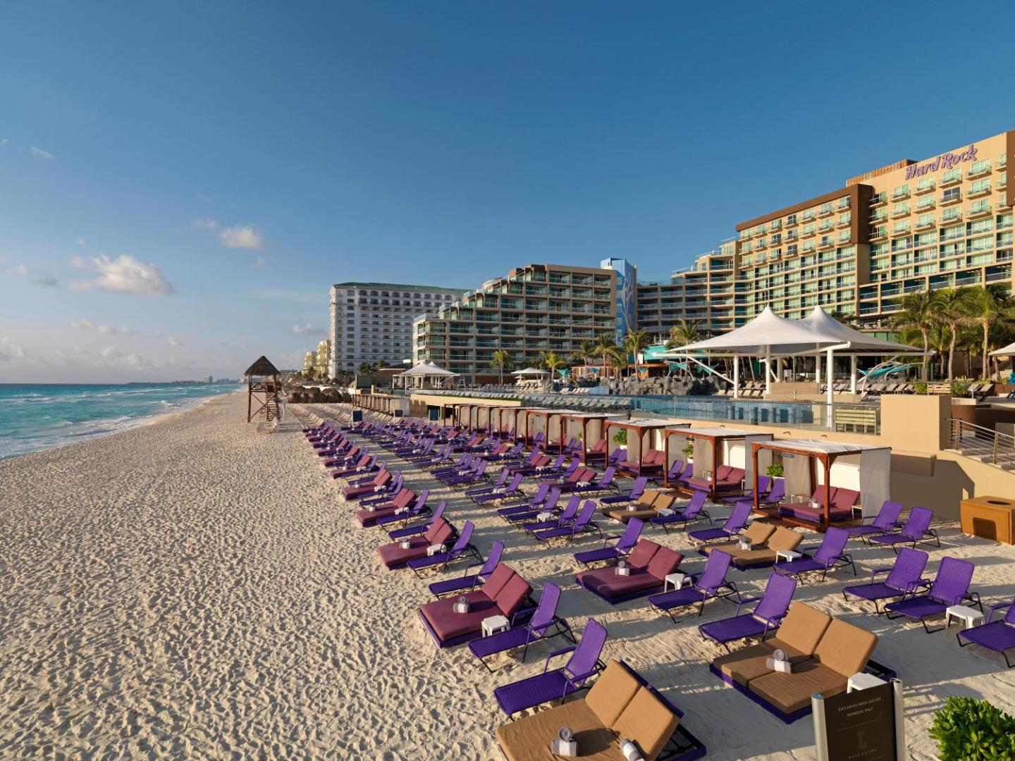 Hard Rock Hotel Cancun privātā pludmale ar violetiem sauļošanās krēsliem, nojumēm un skatu uz viesnīcas kompleksu