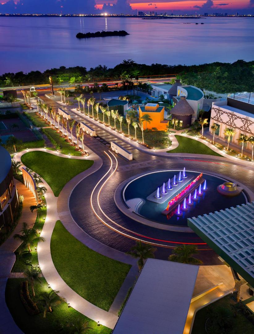 Hard Rock Hotel Cancun teritorijas skats no augšas vakarā ar izgaismotiem celiņiem, palmām un jūras panorāmu Kankunā