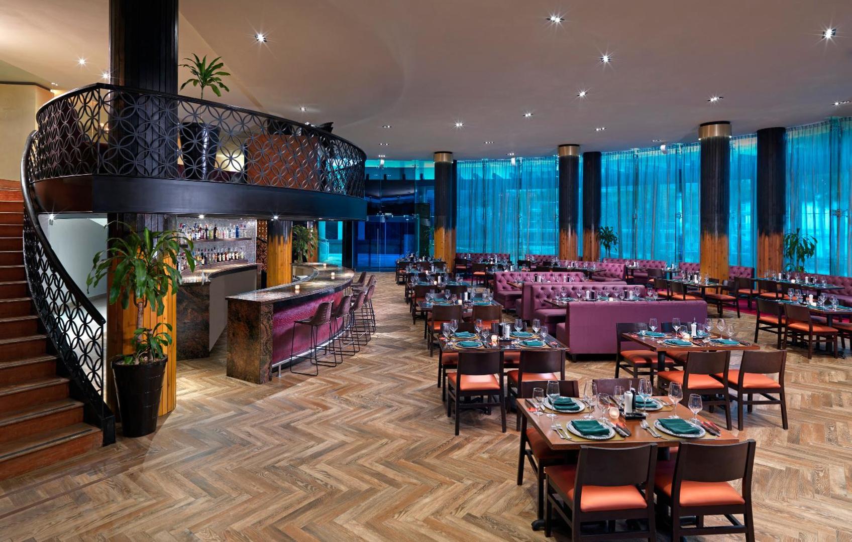 Hard Rock Hotel Cancun restorāna interjers ar bāru, modernām mēbelēm, koka grīdu un elegantiem galdiņu klājumiem