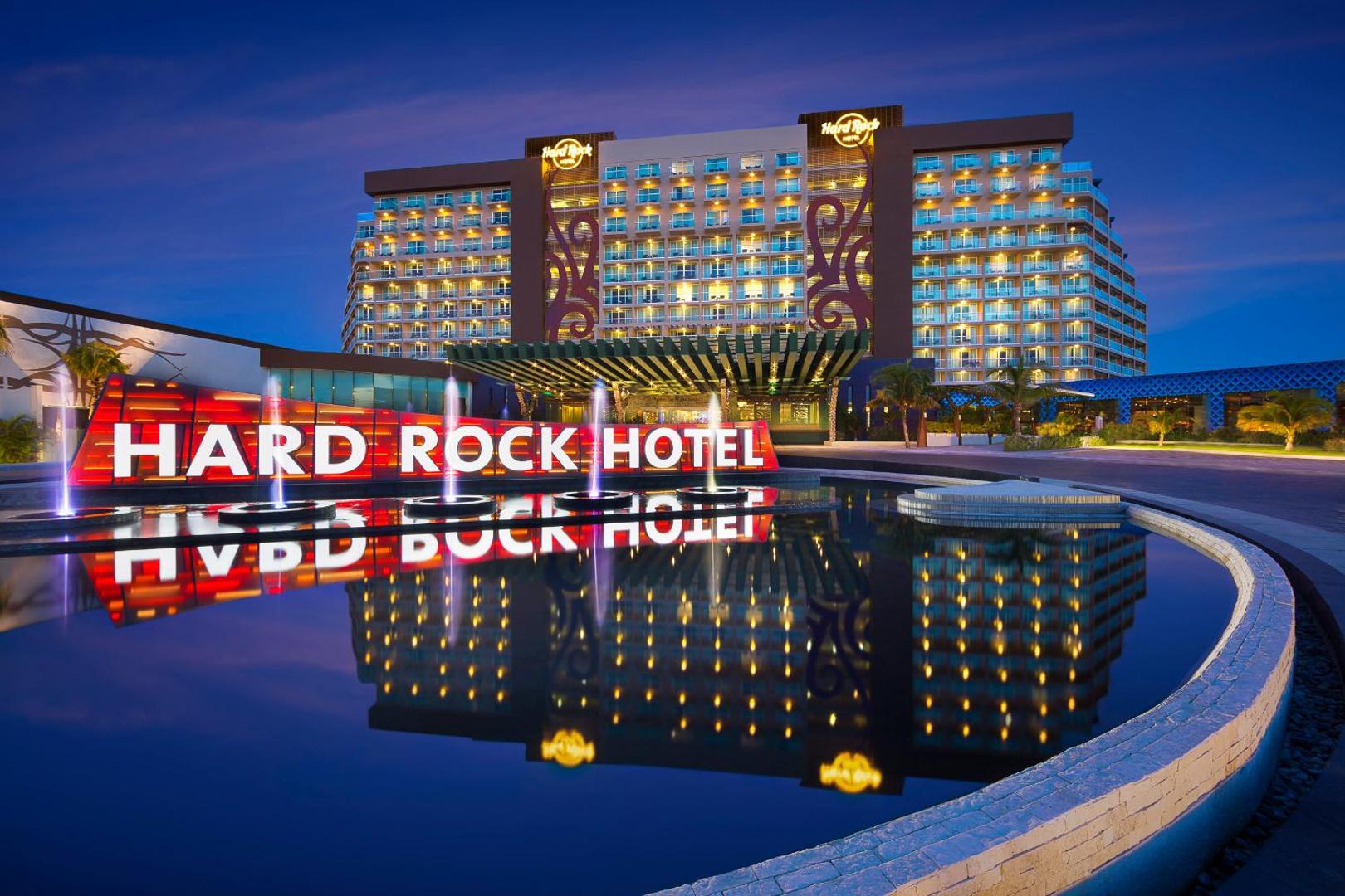 Hard Rock Hotel Cancun fasāde vakarā ar izgaismotu viesnīcas nosaukumu un strūklaku Kankunā, Meksikā — moderna arhitektūra un gaismu spēles