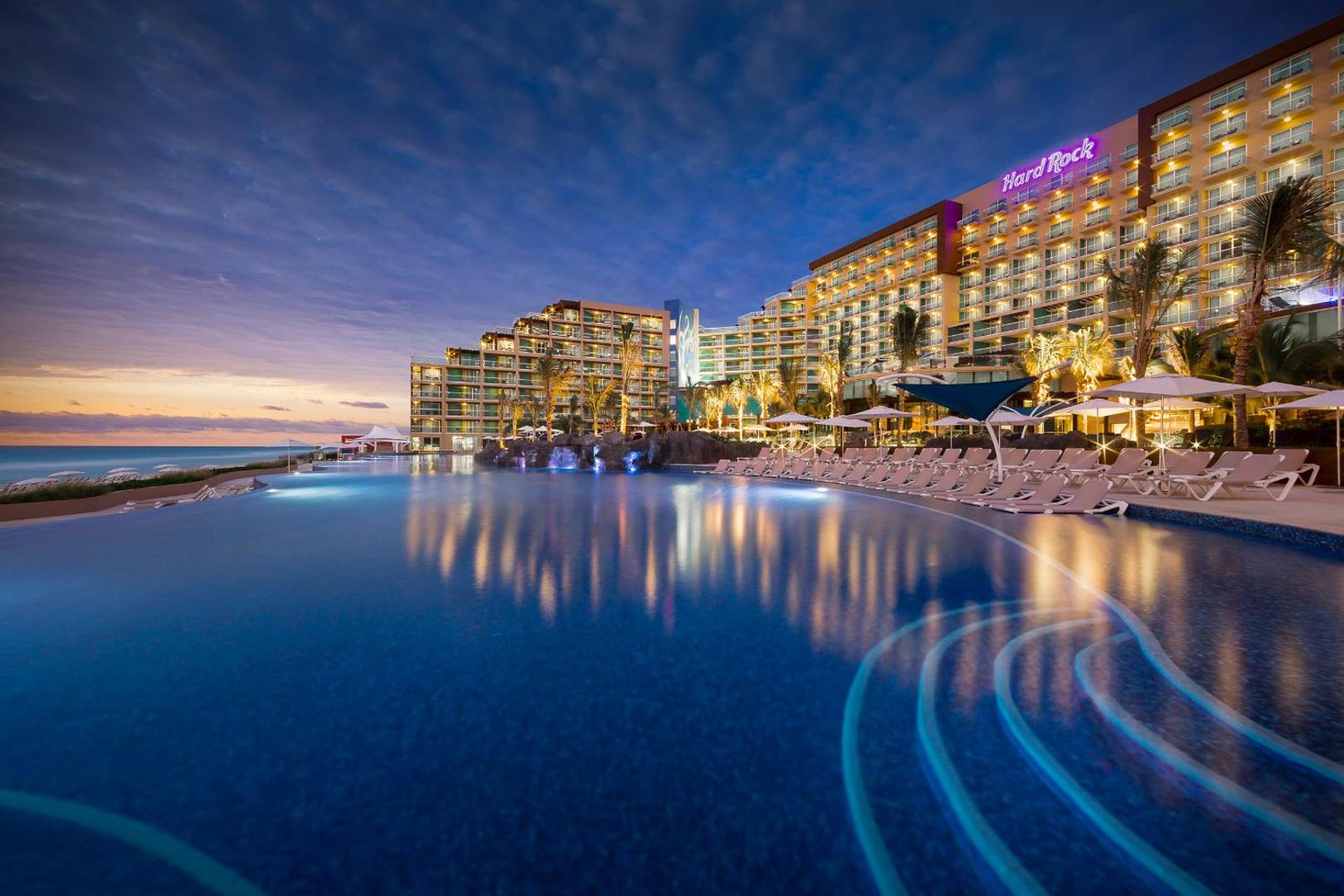 Hard Rock Hotel Cancun āra baseins saulrietā ar apgaismotu viesnīcas ēku, palmām un skatu uz Karību jūru Meksikā