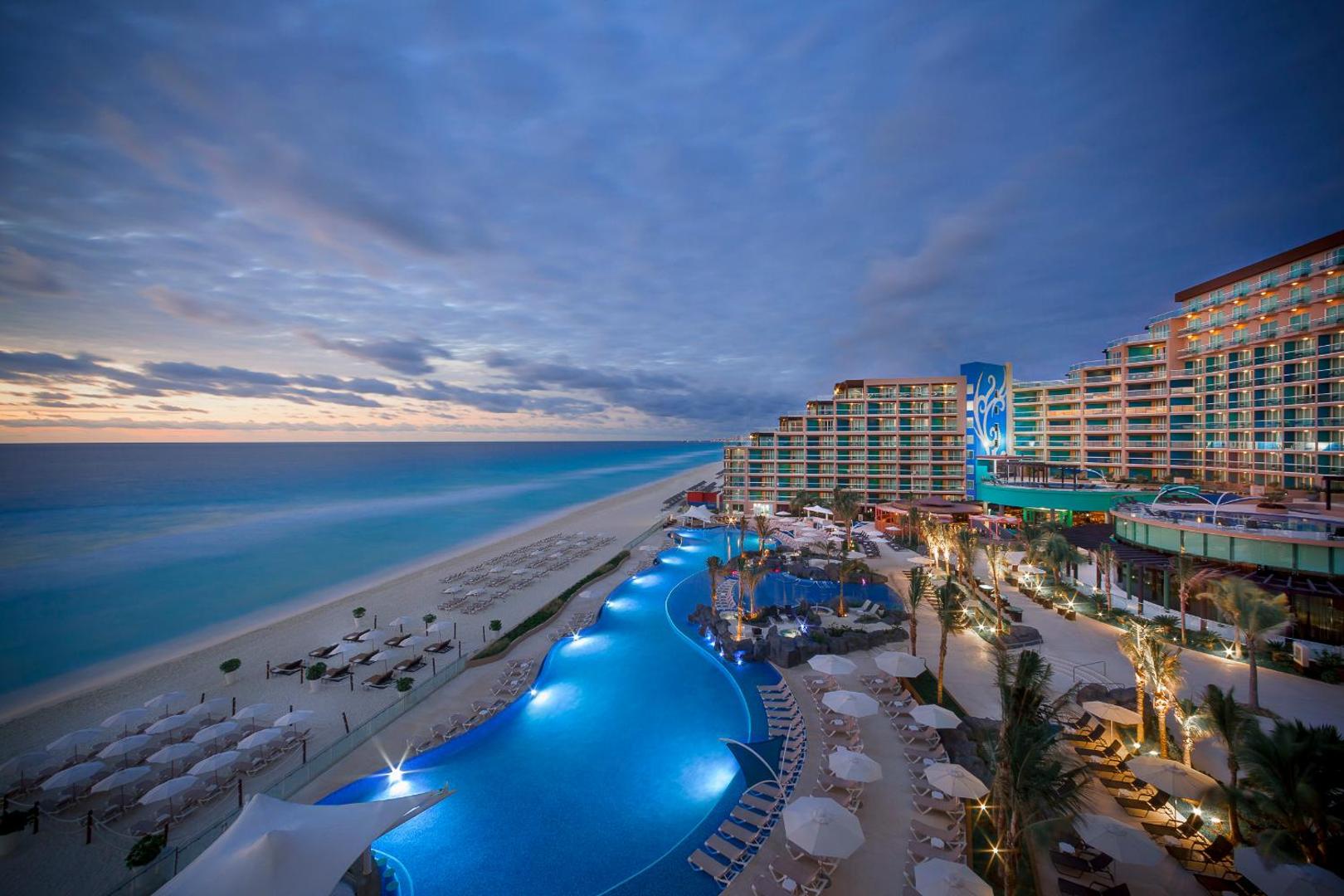 Hard Rock Hotel Cancun panorāmas skats uz baseinu kompleksu, pludmali un viesnīcas ēku vakarā Kankunā