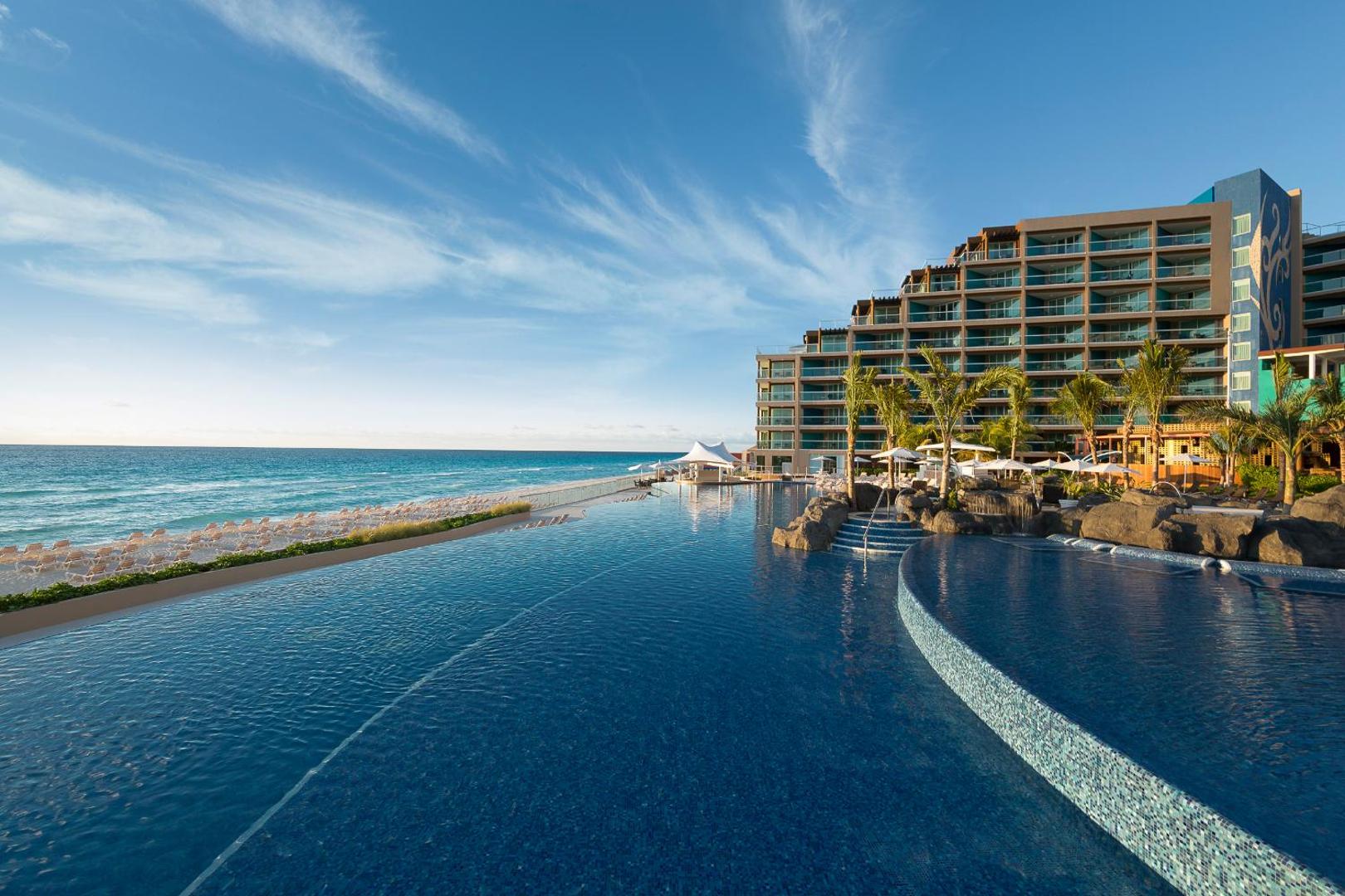 Hard Rock Hotel Cancun bezmalas baseins ar skatu uz tirkīzzilo okeānu un viesnīcas arhitektūru saulainā dienā Kankunā
