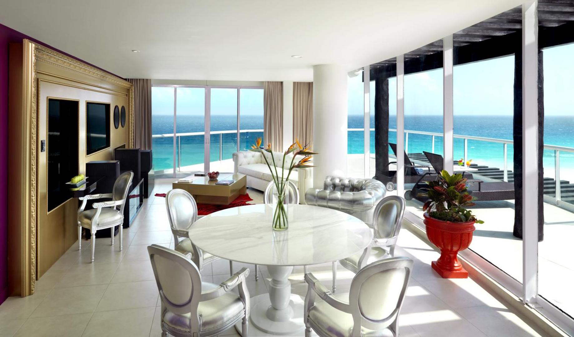 Hard Rock Hotel Cancun luksusa numura viesistaba ar baltām mēbelēm, marmora galdu un panorāmas skatu uz Karību jūru