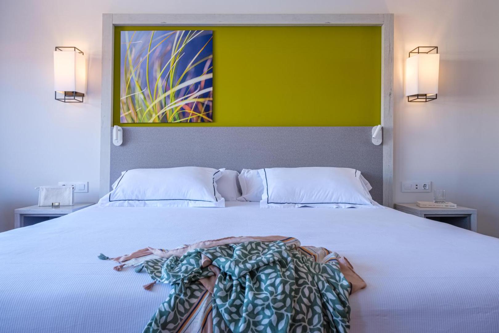 Oassium Hotel numura lielā gulta ar baltu veļu, dekoratīvu paneli zaļā krāsā un moderniem sienas gaismekļiem tuvplānā.