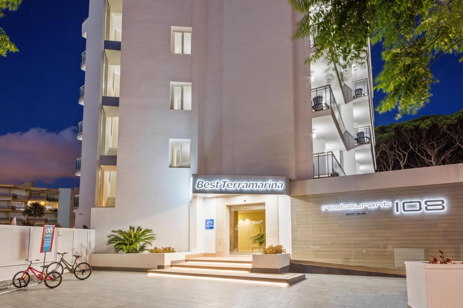 Best Terramarina viesnīcas ieeja un restorāns 108 nakts apgaismojumā — moderna balta fasāde ar velosipēdu novietni priekšplānā.