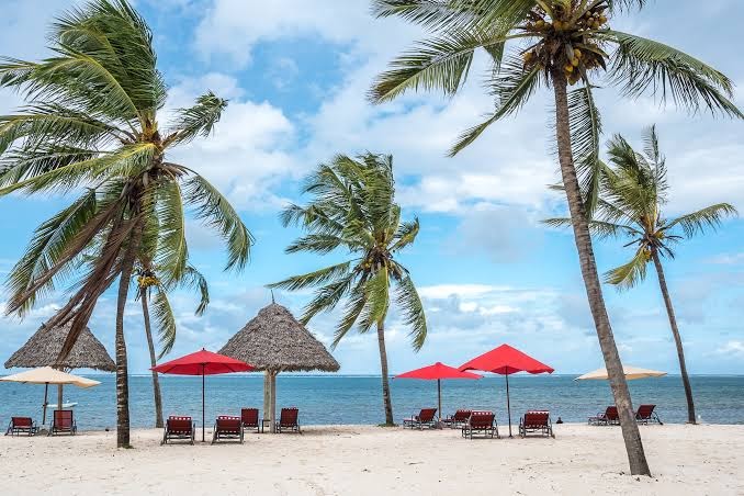 PrideInn Paradise Beach Resort & Spa pludmales atpūtas zona ar sarkaniem saulessargiem un tradicionālām nojumēm Mombasas piekrastē
