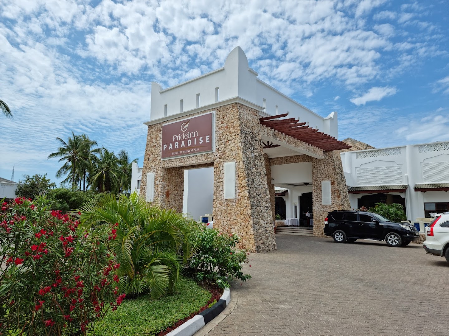 PrideInn Paradise Beach Resort & Spa galvenā ieeja ar viesnīcas logo uz akmens sienas, palmām un bruģētu piebraucamo ceļu