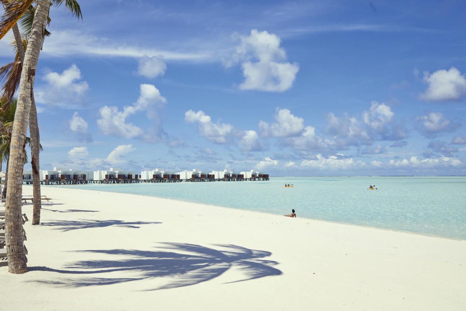 Hotel Riu Atoll baltu smilšu pludmale ar palmām un ūdens bungalo fonā pie tirkīzzila okeāna Maldīvijā
