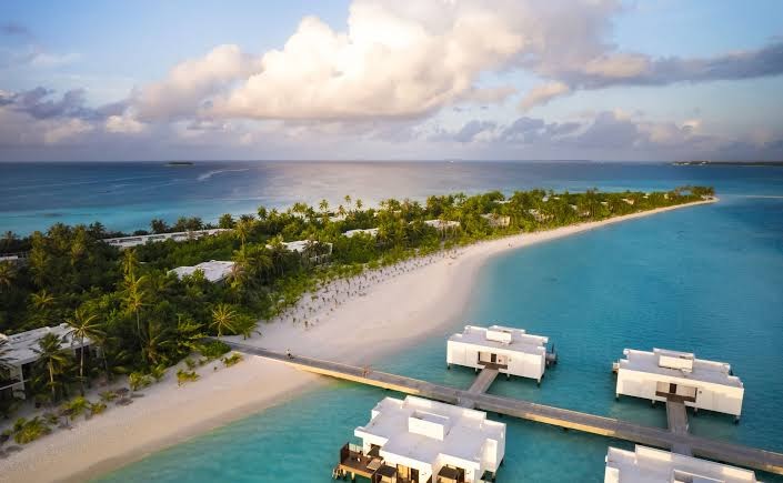 Hotel RIU Atoll ūdens bungalo Maldivu salās saulrieta gaismā — skats uz pludmali, palmām un koka laipām virs dzidrā okeāna ūdens
