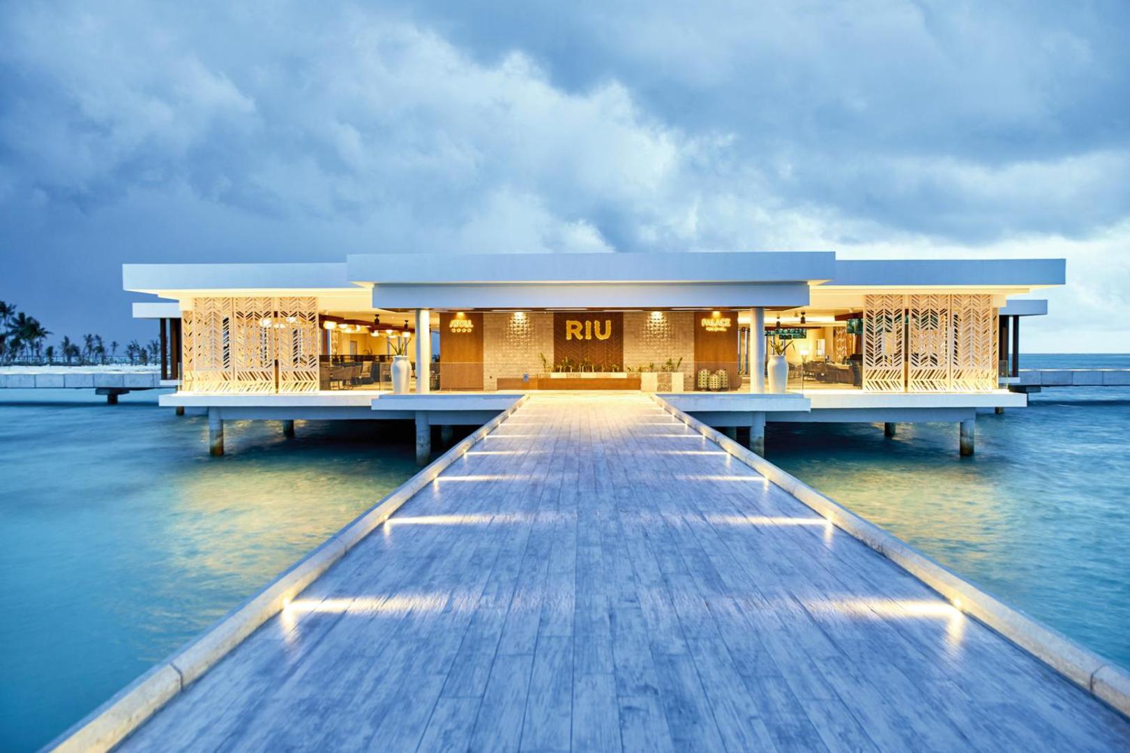 Hotel RIU Atoll galvenā ieeja un vestibils uz pāļiem okeānā — moderna arhitektūra ar izgaismotu koka laipu vakara stundā