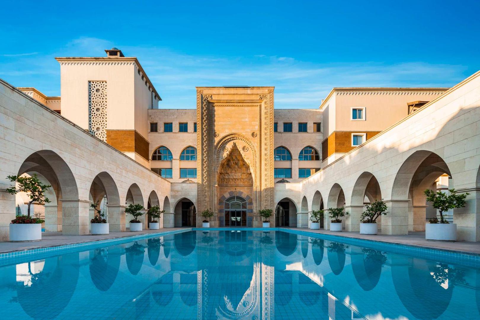 Kempinski Hotel The Dome centrālais pagalms Belekā ar lielu baseinu, ko ieskauj arkādes un senās Anatolijas stila arhitektūras elementi