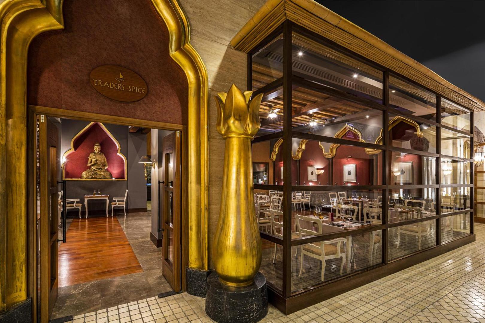 Restorāna Traders Spice ieeja Kempinski Hotel The Dome viesnīcā — zelta krāsas portāls, Budas statuja un stiklota fasāde vakarā
