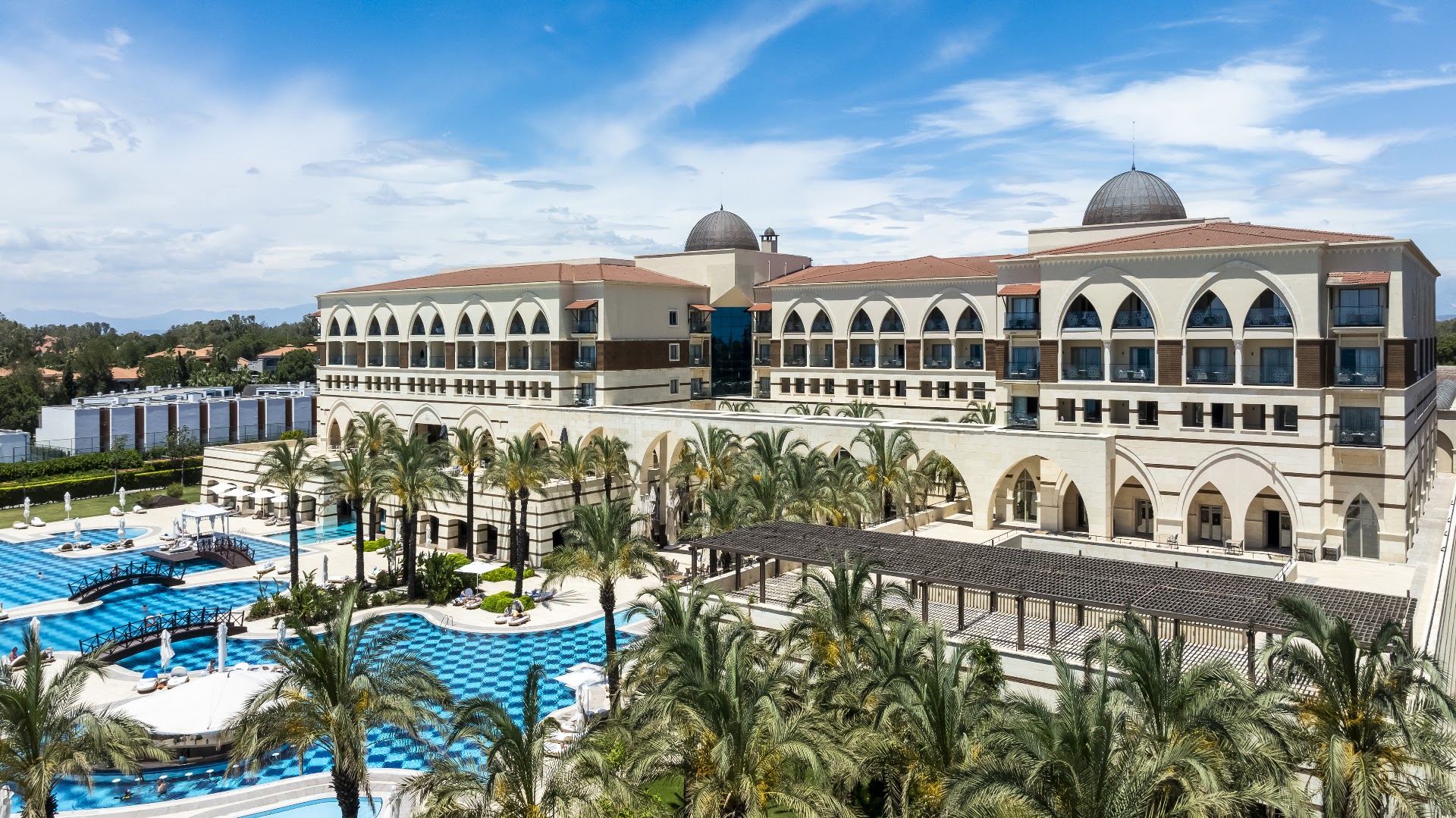 Kempinski Hotel The Dome galvenā ēka ar kupoliem un arkām, lielu āra baseinu un palmām Belekā, Turcijā.