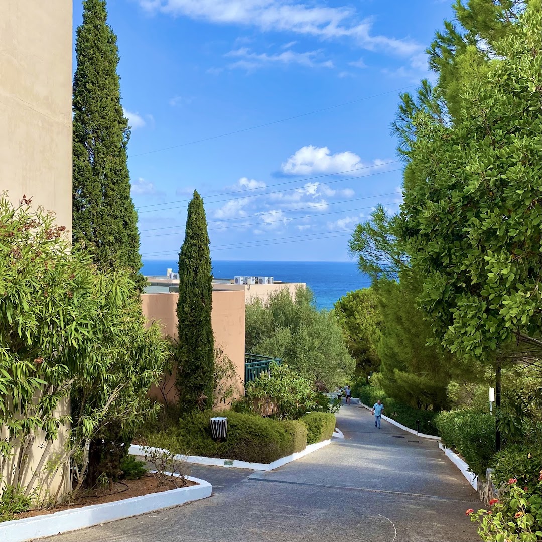 Labiekārtots gājēju ceļš Zeus Hotels The Village Resort & Waterpark teritorijā ar cipresēm un skatu uz jūru tālumā.