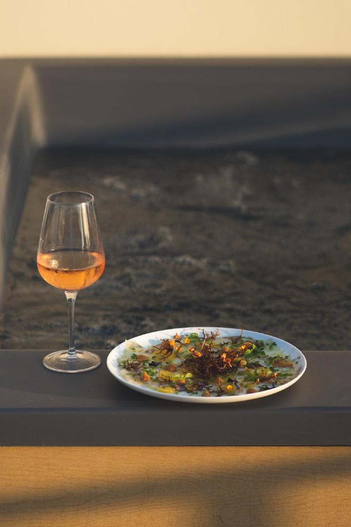 Gourmet uzkoda un glāze rozā vīna uz Zeus Hotels The Village Resort restorāna terases piekrastē saulrieta gaismā.