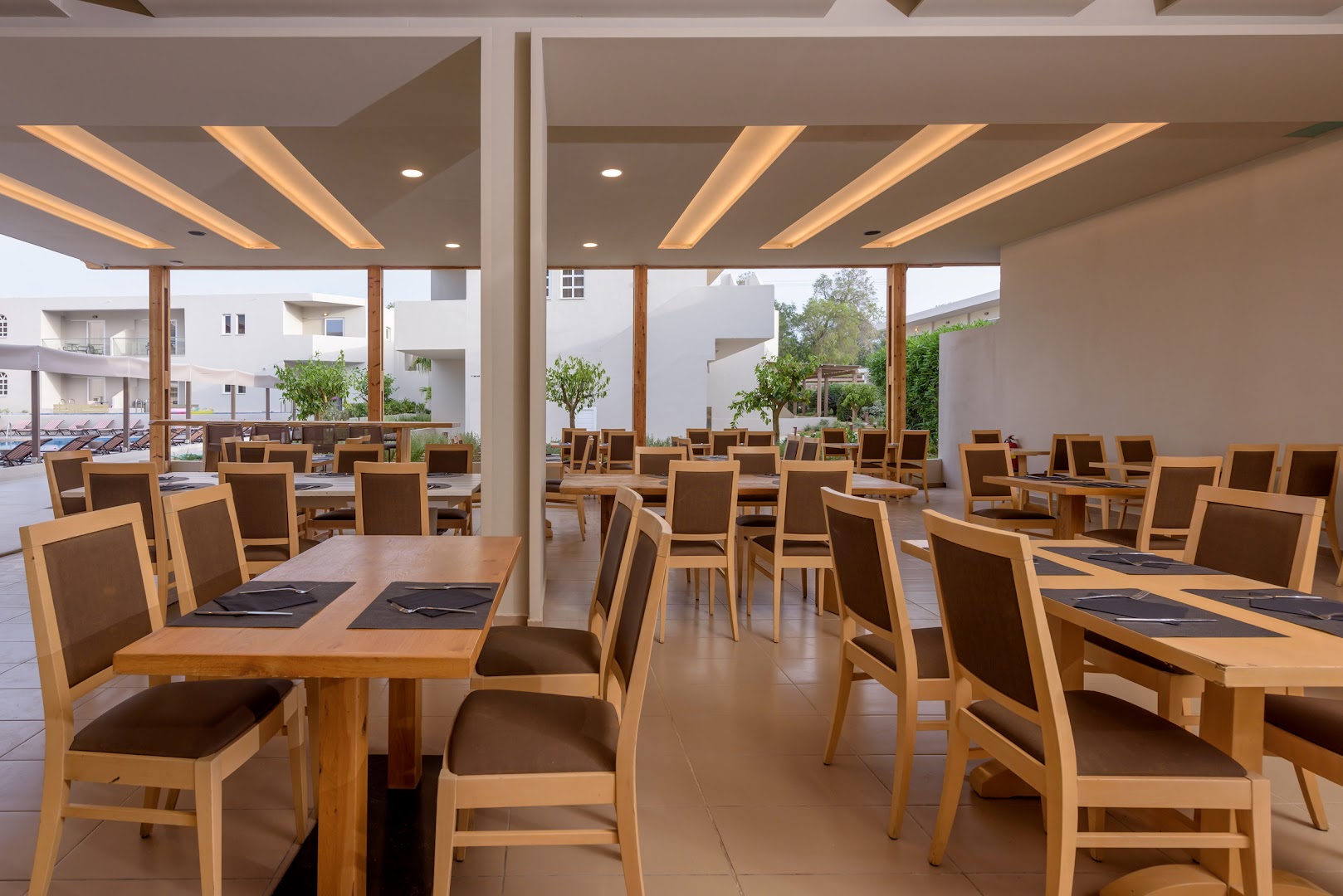 Zeus Hotels Ydoria Resort modernā restorāna zāle ar koka galdiem, stilīgu apgaismojumu un skatu uz viesnīcas iekšpagalmu